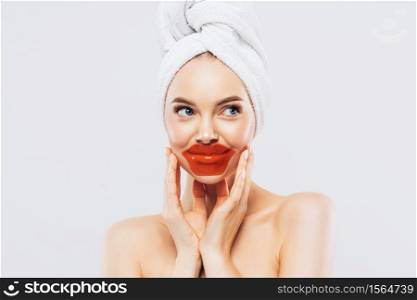 Indoor shot of calm relaxed woman has fresh healthy skin, wears patches on lips, wears white towel on head, gets facial treatment in spa salon