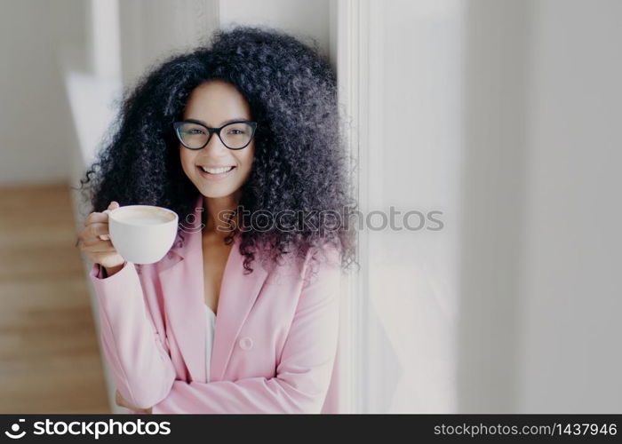 Indoor shot of cheerful curly haired African American lady has coffee break, holds white cup of drink, wears optical spectacles, formal outfit, stands near white wall, works in business sphere