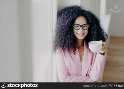 Indoor shot of cheerful curly haired African American lady has coffee break, holds white cup of drink, wears optical spectacles, formal outfit, stands near white wall, works in business sphere