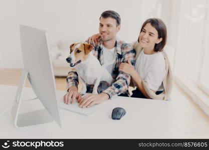 Indoor shot of cheerful husband and wife look with joyful expressions, laugh as watch funny movie, rest together at free time, curious dog looks attentively at monitor of computer, man keyboards