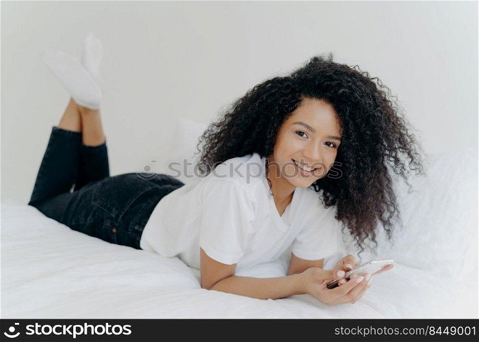 Indoor shot of cheerful millennial woman with Afro hairstyle, lies on stomach in comfortable bed, chats online, checks email box, wears white t shirt, socks and black trousers, has lazy afternoon