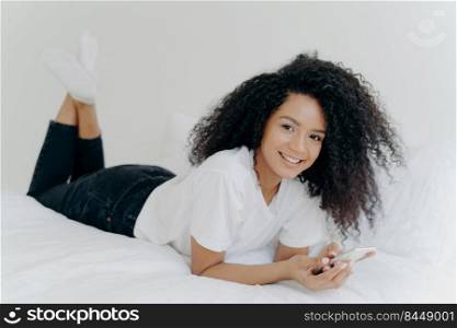 Indoor shot of cheerful millennial woman with Afro hairstyle, lies on stomach in comfortable bed, chats online, checks email box, wears white t shirt, socks and black trousers, has lazy afternoon
