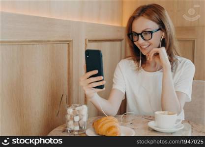 Indoor shot of cheerful redhead young woman uses mobile phone and earphones for online conversation dressed casually sits in cozy coffee shop has friendly chat with best friend being in touch. Indoor shot of cheerful redhead young woman uses mobile phone and earphones for online conversation
