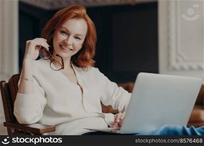 Indoor shot of cheerful satisfied woman works remotely, busy with distance job, looks positively at camera before start working, uses modern laptop computer and wireless internet, browses information