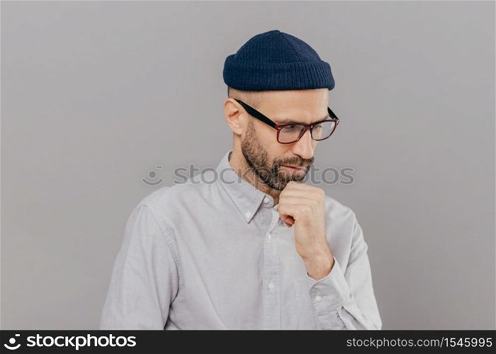 Indoor shot of concentrated man holds chin and focused down, being in deep in thoughts, thinks about innovative idea for his research, has clever expression, wears glasses and white formal shirt