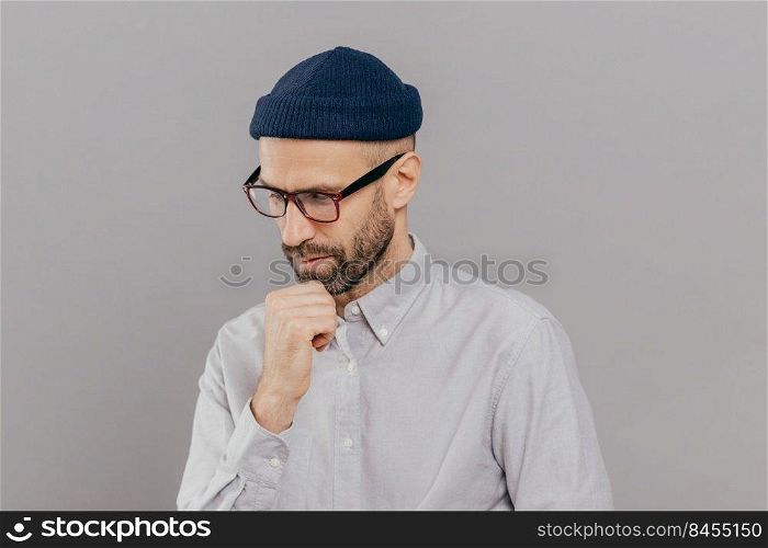 Indoor shot of concentrated man holds chin and focused down, being in deep in thoughts, thinks about innovative idea for his research, has clever expression, wears glasses and white formal shirt