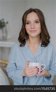 Indoor shot of dreamy young European woman with charming look, dressed in striped pyjamas, drinks coffee, thinks about something, enjoys calm domestic atmosphere. People, drinking and home concept