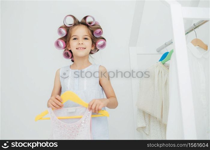 Indoor shot of fashionable cute small girl with gentle warm smile, prepares for something, has curlers on hair, holds new outfit on hangers, look joyfully at camera. Children and beauty concept