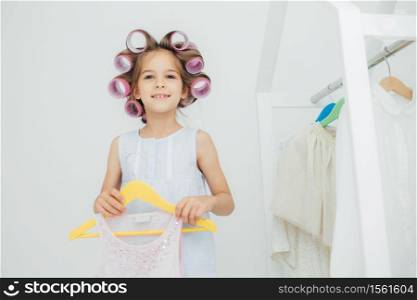 Indoor shot of fashionable cute small girl with gentle warm smile, prepares for something, has curlers on hair, holds new outfit on hangers, look joyfully at camera. Children and beauty concept