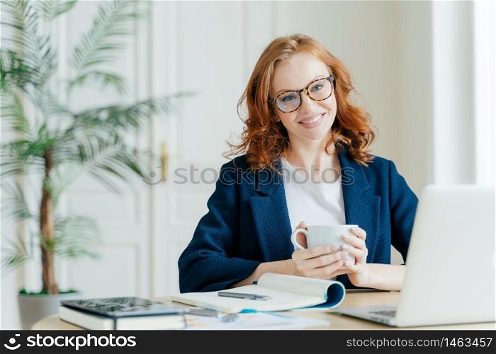 Indoor shot of female editor works on article, uses laptop computer and wireless internet, checks latest news, holds mug of coffee or tea, surrounded with notepad for writing notes, smiles happily