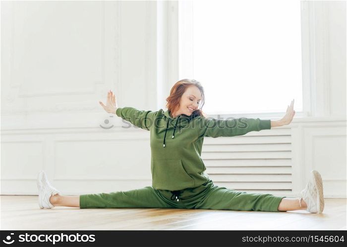 Indoor shot of fit female stretches hand and legs sideways, sits in pose of splits, tilts head on right, has toothy smile, red hair, wears tracksuit, poses in studio, engaged in yoga. Flexibility