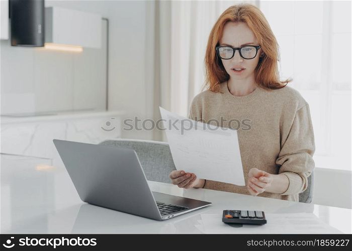 Indoor shot of ginger professional female economist calculates finance at home holds paper document makes financial report sits at white desktop surrounded by modern laptop computer and calculator. Professional female economist calculates finance at home holds paper document makes financial report