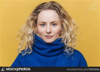 Indoor shot of good looking of serious blue eyed female wears blue warm sweater, looks confidently at camera, isolated over yellow background. Studio shot of beautiful curly woman models in studio
