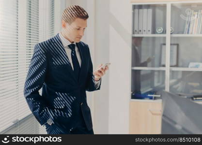 Indoor shot of handsome male business owner, reads text message, stands near office window, poses in office, has concentrated look in smart phone. Director checks financial report in database