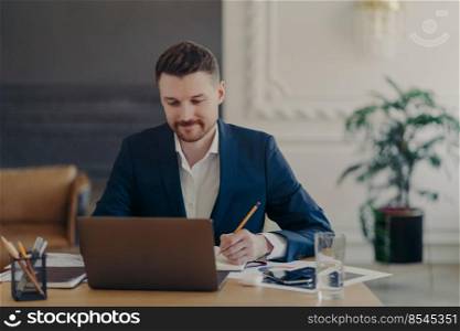 Indoor shot of handsome male office worker rewrites information from notepad in diary sits at desk dressed in formal clothes spends time for searching info. Businessman takes notes does paper work