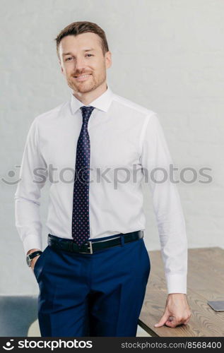 Indoor shot of handsome unshaven male entrepreneur wears formal shirt, tie and trousers, keeps hand in pocket, meets with business partners. Prosperous young businessman rejoices his success