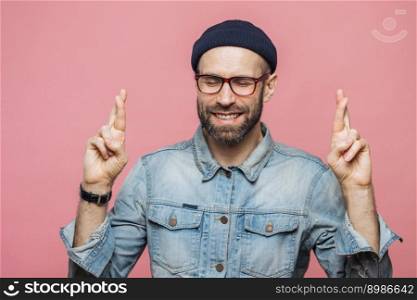 Indoor shot of handsome unshaven male keeps fingers crossed, eyes shut, has happy expression, dressed in fashionable clothing isolated over pink background. Man hopes for better before important event