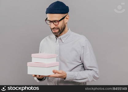 Indoor shot of handsome unshaven man with brislte holds two boxes, happy to recieve present from friend on birthday, wears festive clothes, models against grey background. Young male with gift