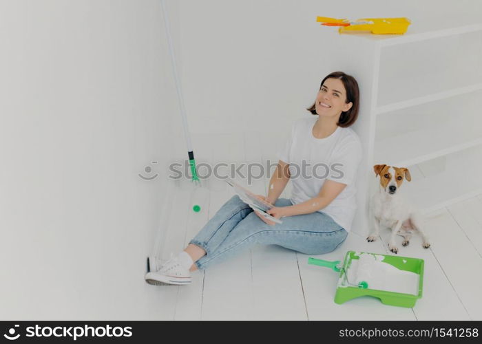 Indoor shot of happy brunette woman in casual wear, sits on floor near white drawer, holds color samples, her jack russell terrier dog poses near, busy with refurbishing walls in bought house