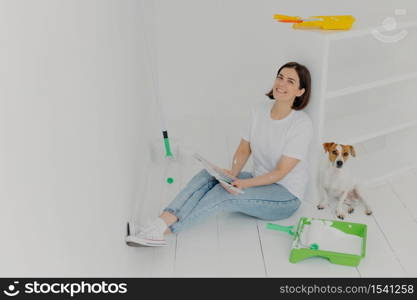 Indoor shot of happy brunette woman in casual wear, sits on floor near white drawer, holds color samples, her jack russell terrier dog poses near, busy with refurbishing walls in bought house