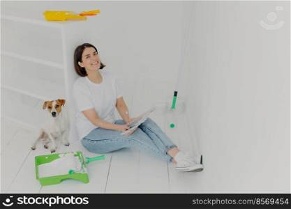 Indoor shot of happy brunette woman in casual wear, sits on floor near white drawer, holds color s&les, her jack russell terrier dog poses near, busy with refurbishing walls in bought house
