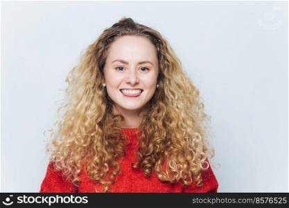 Indoor shot of happy curly blonde woman with pleasant smile and cheerful expression, dressed in warm red sweater, models against white background. People, happiness and positive emotions concept
