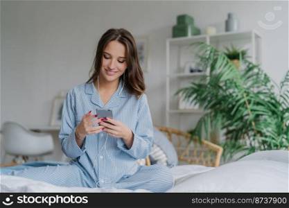 Indoor shot of happy dark haired woman in casual domestic costume, enjoys rest time, poses in cozy bedroom, enjoys music with modern devices, domestic interior in background. People, lifestyle concept