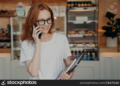 Indoor shot of happy ginger shop owner has telephone conversation holds mobile phone near ear carries notepad and laptop computer satisfied with tariffs of call wears spectacles white t shirt. Happy ginger shop owner has telephone conversation holds mobile phone near ear