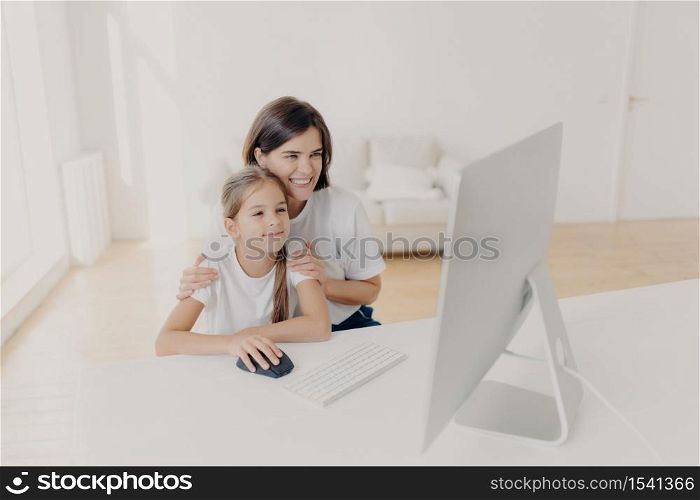 Indoor shot of happy lovely mother and daughter watch funny cartoon on computer, have pleasant smiles, enjoy togetherness, spend free time or weekend at home, spacious light empty living room