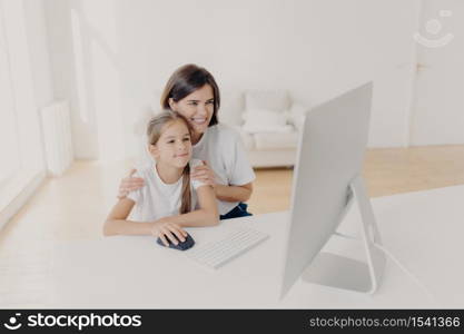 Indoor shot of happy lovely mother and daughter watch funny cartoon on computer, have pleasant smiles, enjoy togetherness, spend free time or weekend at home, spacious light empty living room