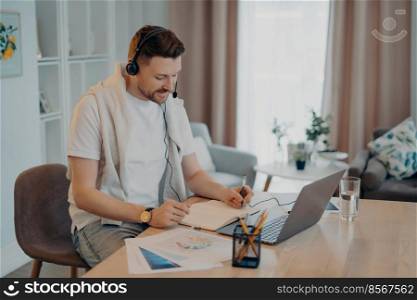 Indoor shot of happy male makes notes while listens online courses studies remotely from home sits at desktop with modern laptop computer papers wears headphones has web conference or virtual meeting