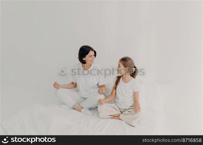 Indoor shot of happy mom and daughter have pleasant talk together, have yoga practice, meditate on soft bed, dressed casually, pose on white bedclothes, have good relationships. Morning relaxation