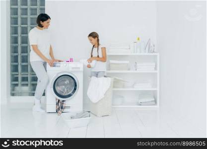 Indoor shot of happy mother and daughter stand near washing machine, girl pours liquid powder, load washer with dirty clothes, do housework, have laundry day at home. Household chores concept