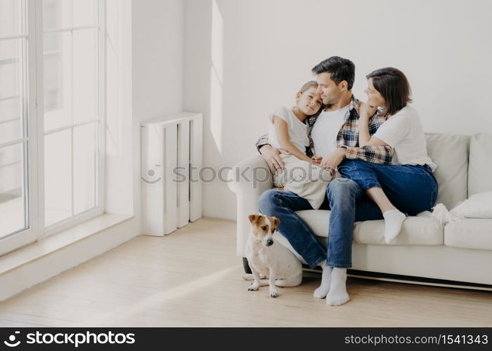 Indoor shot of happy mother and father spend weekend with small daughter, pose all together on comfortable sofa, have fun and pleasant talk with each other. Little dog sits near on floor. Family