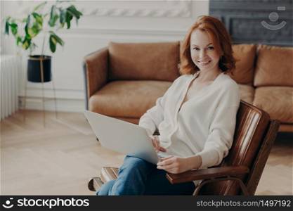 Indoor shot of happy redhead woman freelancer sits at home interior, uses modern laptop computer for distance work, enjoys online communication and free internet connection, waits for answer