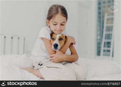 Indoor shot of happy small child embraces favourite pet, dressed in pyjamas, expresses love to her dog, plays with animal after awakening, poses on soft bed. Children and domestic animals concept