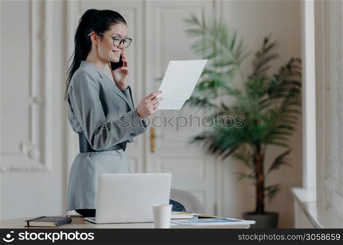 Indoor shot of happy smiling woman manager checks information from papers wears grey formal costume, optical eyewear concentrated on research work has telephone conversation, stands in coworking space