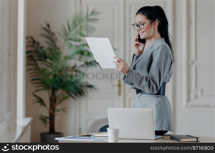 Indoor shot of happy smiling woman manager checks information from papers wears grey formal costume, optical eyewear concentrated on research work has telephone conversation, stands in coworking space