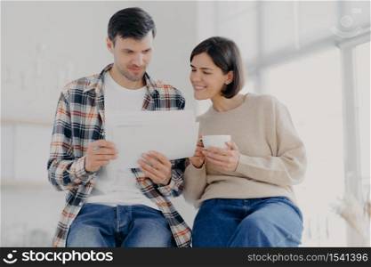 Indoor shot of happy wife and husband study documents received from bank, do paperwork together, drinks aromatic coffee, stand closely to each other, dressed in casual wear, pose in cabinet.