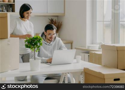 Indoor shot of happy woman and man spend morning at their new kitchen with big windows and modern furniture, husband works on laptop computer discusses with wife trendy design, surrounded with boxes
