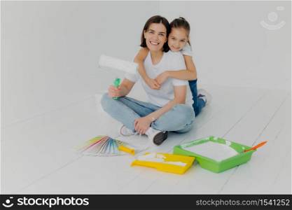 Indoor shot of happy woman gets warm hug from lovely daughter, pose together in empty room, choose bright color for painting from sample palette, busy with house renovation ande redecoration.