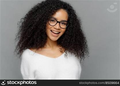 Indoor shot of happy young lady has Afro hairstyle, smiles broadly, glad to be promoted, wears optical glasses and white sweater, models over grey background. Ethnicity, emotions, pleasure concept