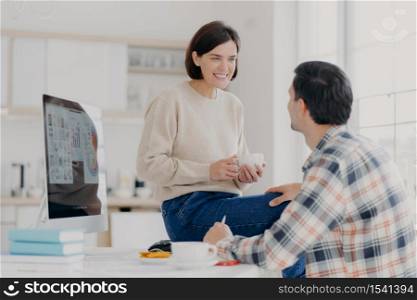 Indoor shot of happy young woman and man discuss something with drink, use modern computer for making financial report, pose in spacious light room, collaborate together for making common task