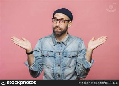 Indoor shot of hesitant bearded man shrugs shoulders, looks uncertain, doubts when makes decision, isolated over pink background. People, doubt and hesitation concept. Body language concept.