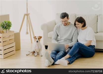 Indoor shot of lovely couple move in new dwelling, use modern laptop for searching design ideas for their flat, sits on floor near sofa, pedigree dog poses near cardboard boxes. Moving Day concept