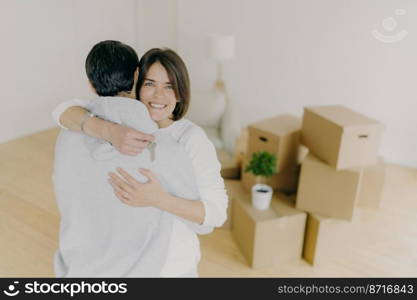 Indoor shot of lovely family couple embrace with love, hold keys from new apartment, move in own apartment, pose in empty light living room with floor l&and pile of many cardboard containers