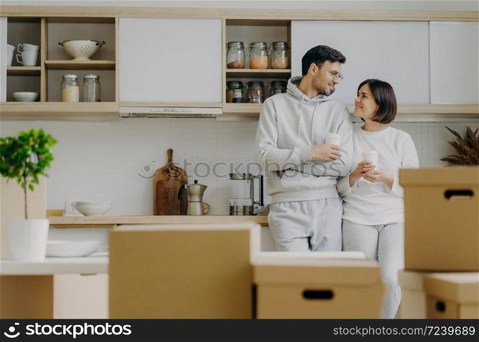 Indoor shot of lovely newlywed couple talk to each other with pleased expressions, drink takeaway coffee from paper cups, pose in modern kitchen with new bought furniture, change place of living