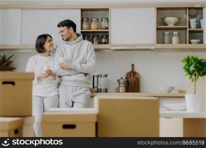 Indoor shot of lovely newlywed couple talk to each other with pleased expressions, drink takeaway coffee from paper cups, pose in modern kitchen with new bought furniture, change place of living