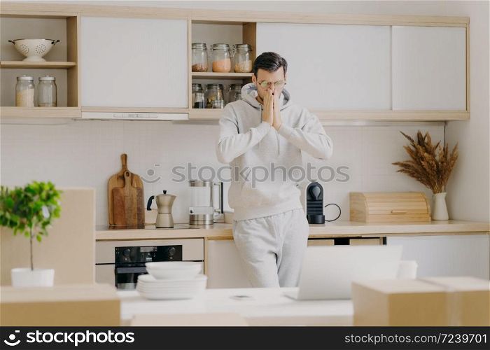 Indoor shot of pensive man works on family budget, looks attentively at laptop computer, poses in kitchen with unpacked boxes, rents new house, dressed in casual sweatshirt, works remotely at home