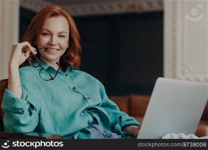 Indoor shot of pleasant looking redhead woman has good time at home, works on computer, wears casual shirt, holds eyeglasses, feels comfort, enjoys domestic atmosphere. Teacher teaches by video call